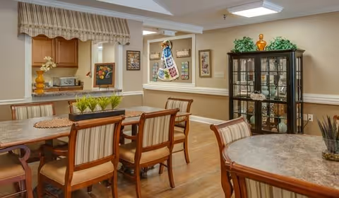 Bright communal dining area with wooden tables and upholstered chairs, a serving pass to a kitchen, and a glass-front china cabinet.