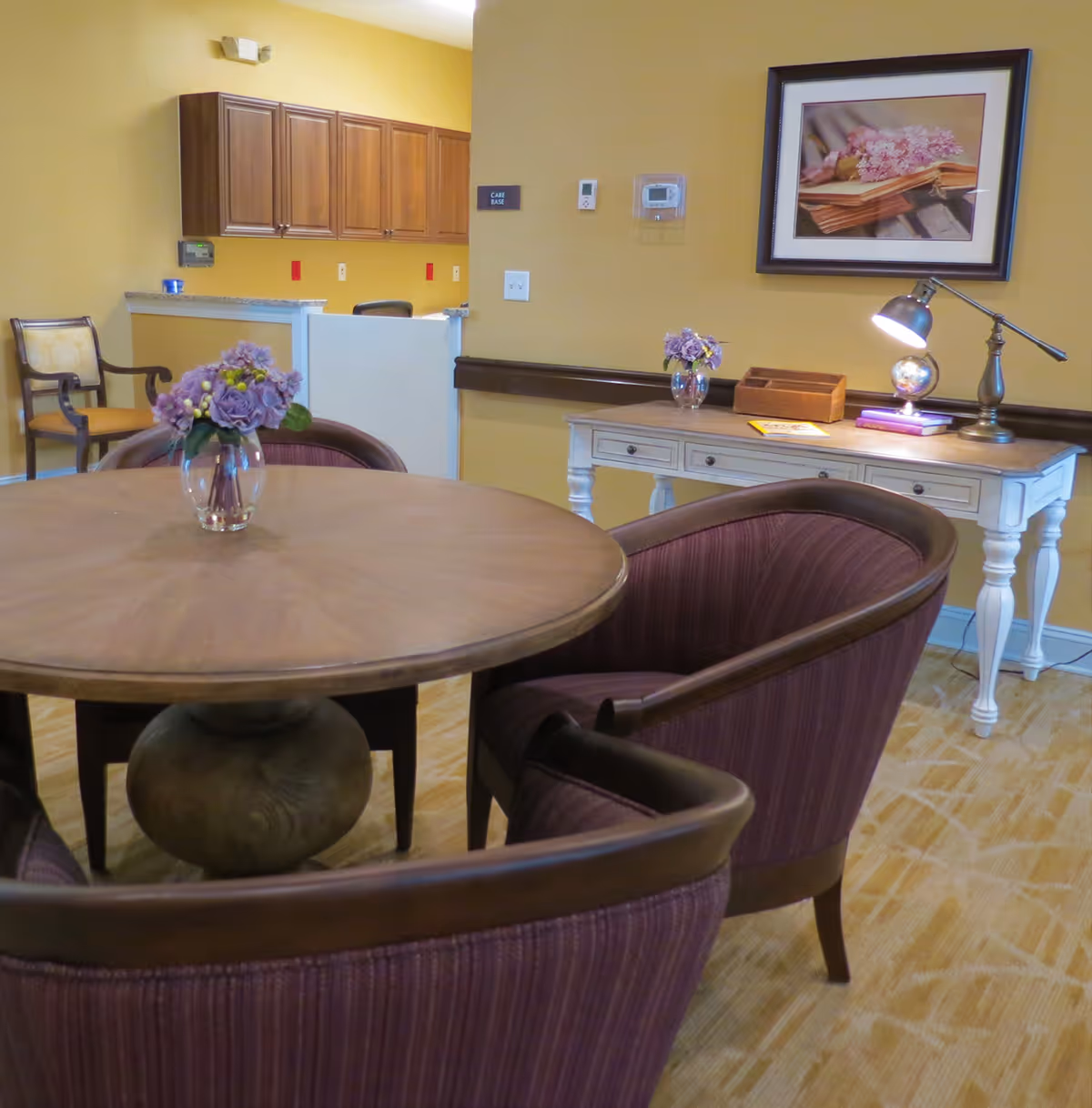 A cozy interior room with a round wooden table surrounded by four purple upholstered chairs. A small glass vase with purple flowers is placed on the table. In the background, there is a white desk with a lamp, books, and another small vase with purple flowers. Above the desk hangs a framed picture of flowers on a book. Wooden cabinets and a chair are visible further back against a yellow wall.