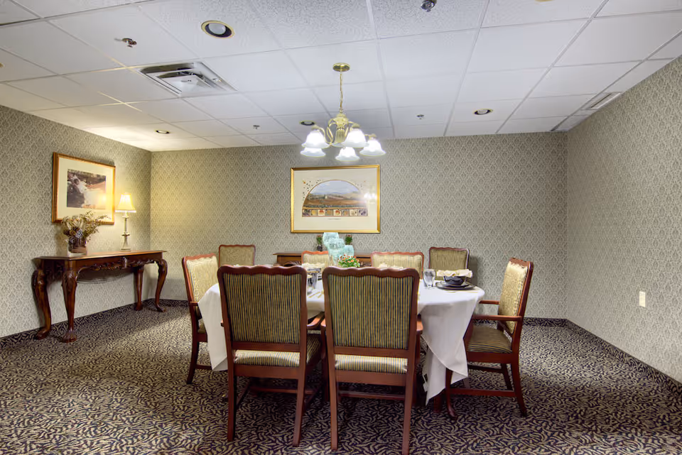 A formal dining room with a round table set for six, upholstered chairs, a sideboard, framed artwork, and an overhead chandelier.