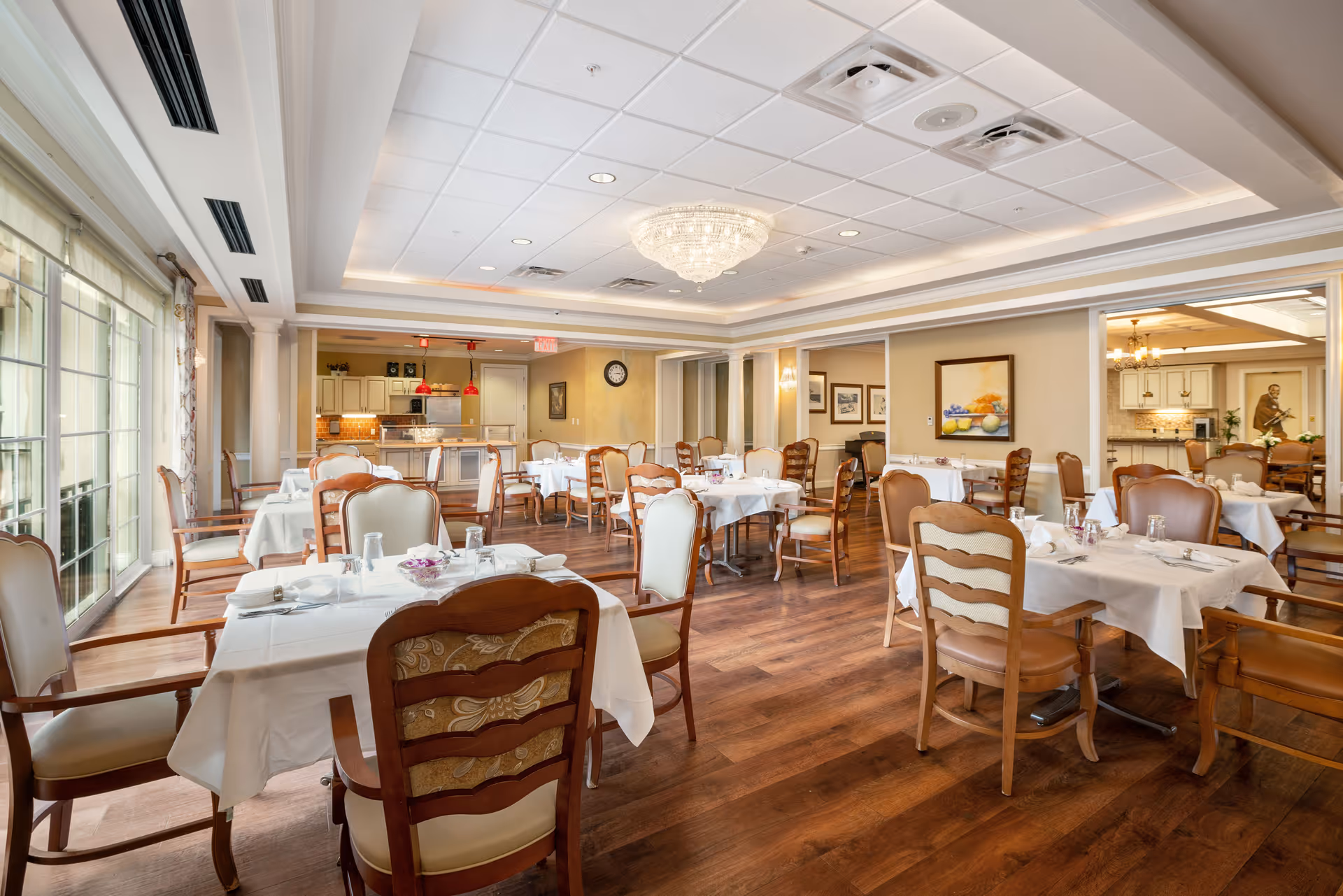 A bright and spacious dining room with multiple tables covered in white tablecloths, set with glasses and napkins. The room features wooden chairs with cushioned seats, large windows with curtains allowing natural light, a chandelier hanging from the ceiling, and a kitchen area visible in the background.