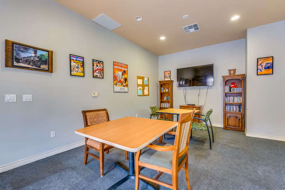 A senior living facility common room with two tables and several chairs arranged around them. The walls are decorated with vintage movie posters and a framed picture of a steam locomotive. There is a large flat-screen TV mounted on the wall, flanked by two wooden bookshelves filled with DVDs and books. The room has carpeted flooring and recessed ceiling lights.