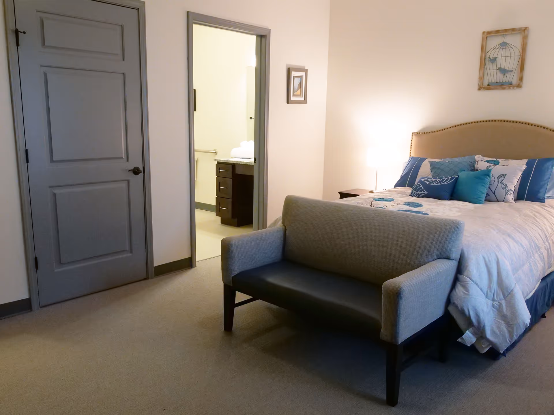Bedroom featuring a made bed with decorative pillows, a small upholstered bench at the foot, a nightstand with lamp, and an open doorway to a bathroom.