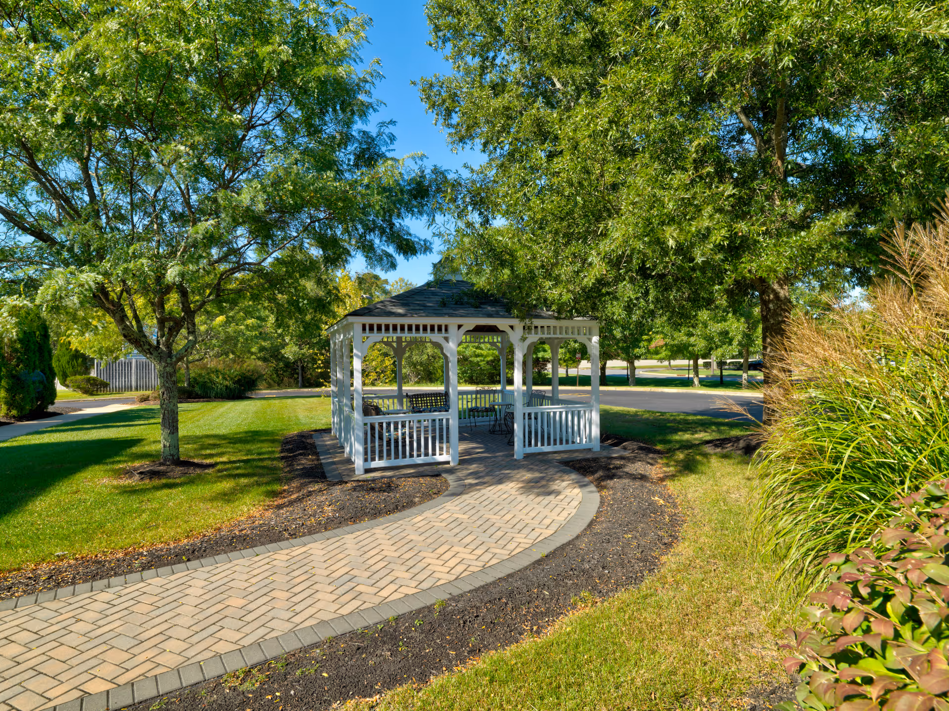 White wooden gazebo at the end of a curved brick walkway surrounded by green trees and lawn.