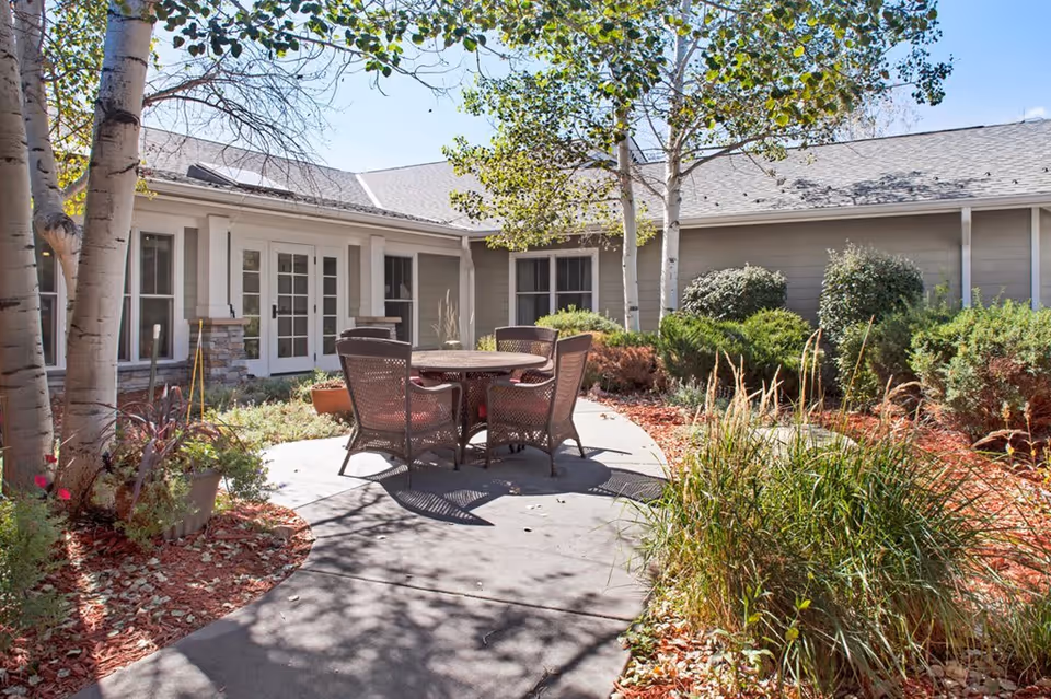 Outdoor patio area at Sunrise of Boulder featuring a round table with four wicker chairs surrounded by trees, shrubs, and landscaped garden beds with mulch and plants under a clear sky.
