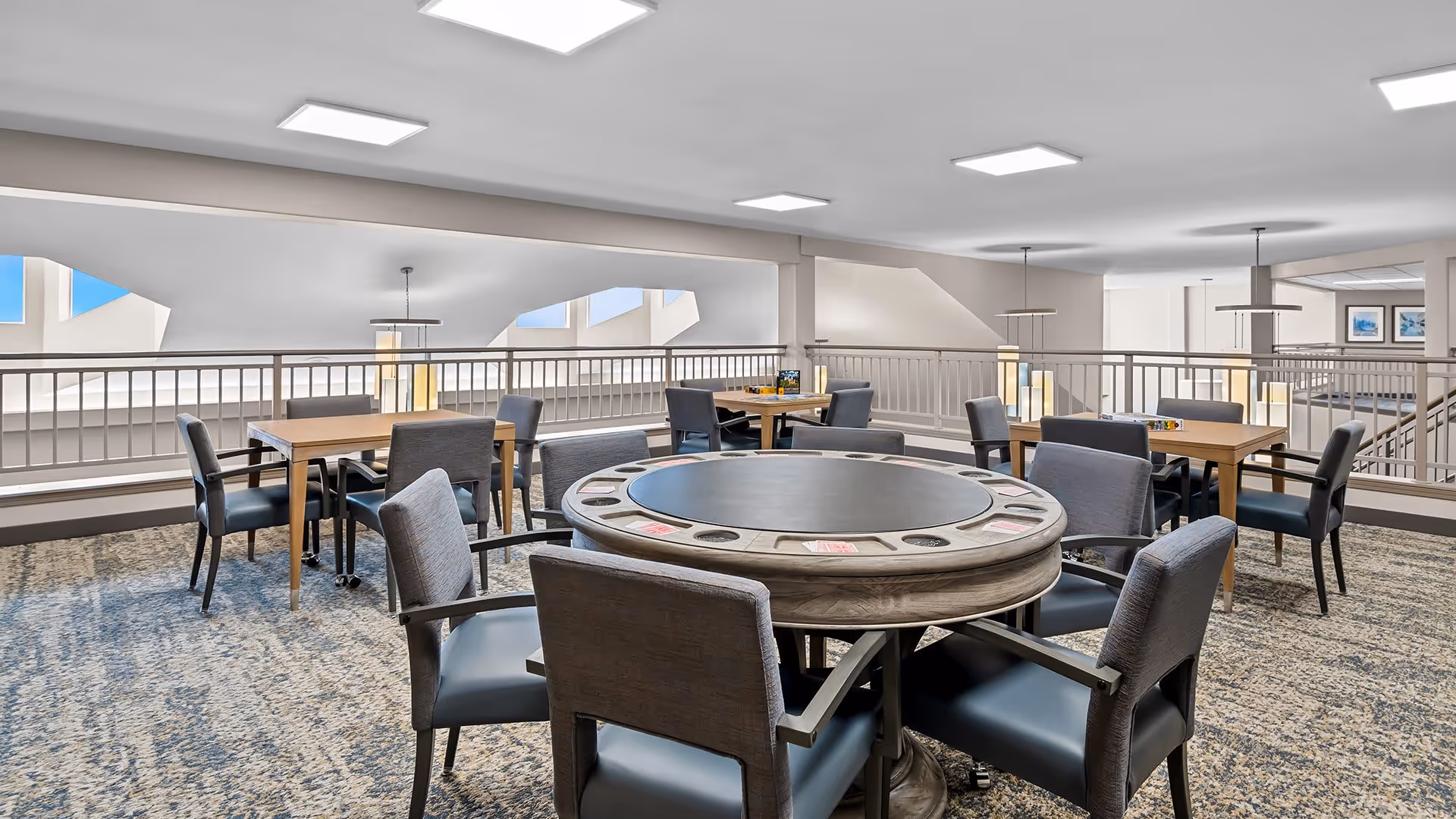 A spacious common area with multiple tables and chairs arranged for group activities or dining. The central table is round with built-in cup holders and card slots, surrounded by cushioned chairs. The room has a carpeted floor, modern ceiling lights, and large windows letting in natural light. Railings separate this upper-level space from the lower floor.