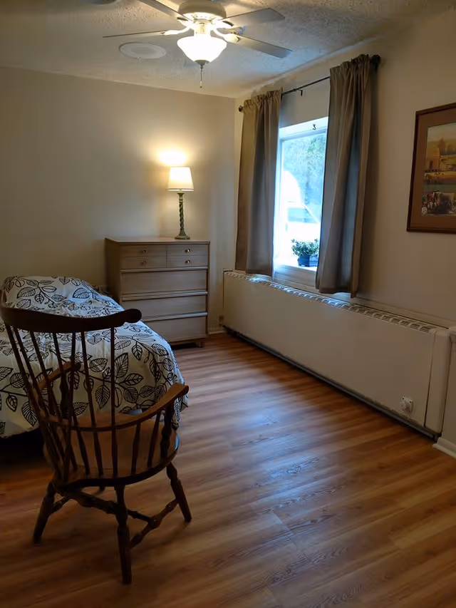 A simple bedroom with a wooden floor, a single bed with a leaf-patterned bedspread, a wooden chair, a wooden dresser with a lit lamp on top, a window with beige curtains, and a framed picture on the wall.