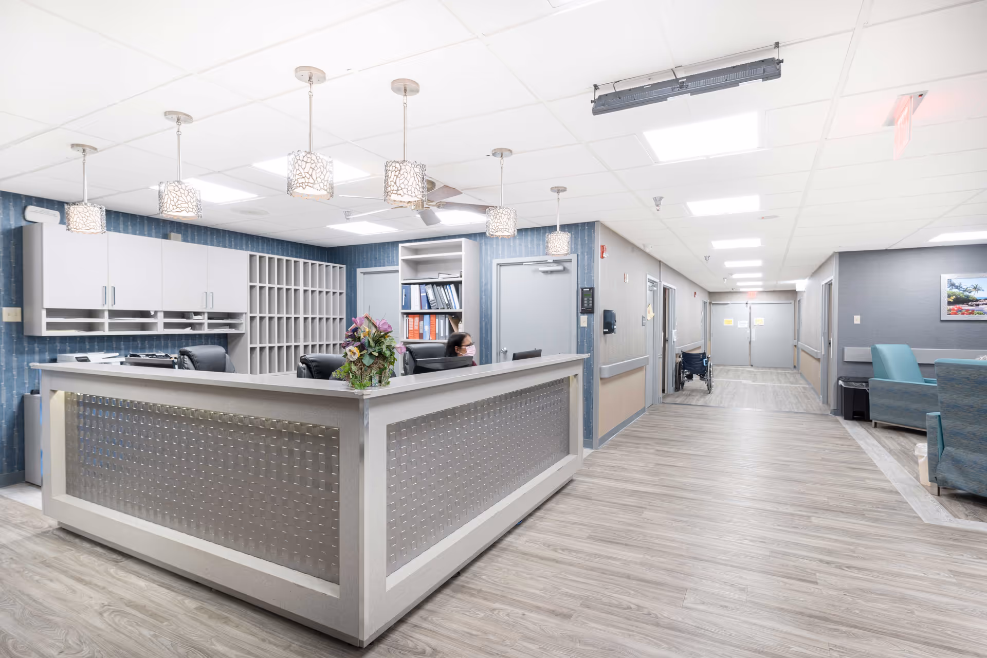 Reception area inside Grandview Nursing & Rehabilitation facility with a front desk, a receptionist wearing a mask, pendant lights hanging from the ceiling, a hallway with doors, a wheelchair, and seating area with blue chairs.