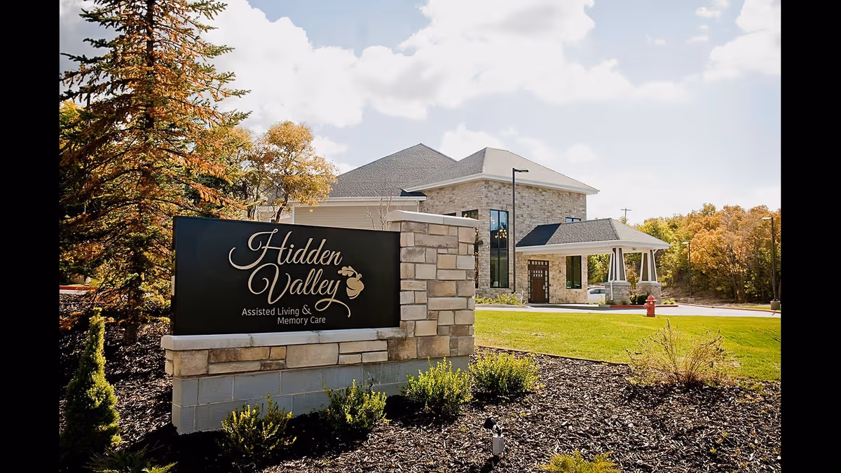 Exterior view of Hidden Valley Assisted Living and Memory Care facility with a stone sign in the foreground displaying the facility's name, surrounded by landscaped greenery and trees under a partly cloudy sky.