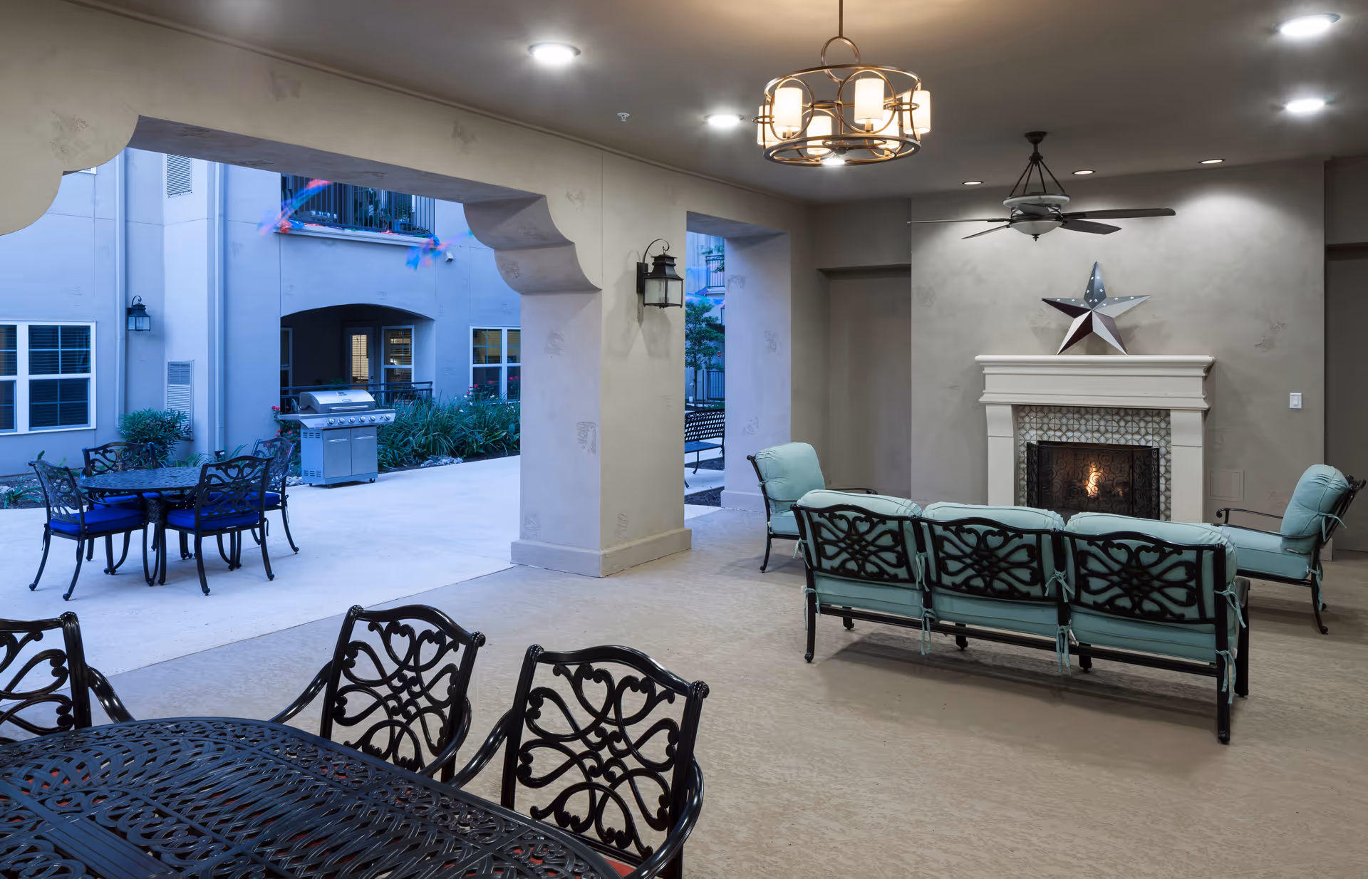 Covered outdoor seating area with cushioned metal chairs and a sofa arranged around a lit fireplace with a star decoration above it. There is a ceiling fan and a chandelier overhead. Beyond the covered area, there is an open patio with a metal dining table and chairs, a barbecue grill, and surrounding plants and building walls.