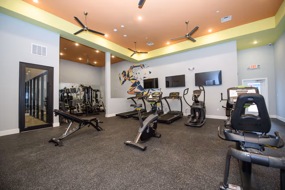 Interior view of a fitness room in Hamilton Parc facility featuring exercise equipment including stationary bikes, treadmills, elliptical machines, and a weight bench. The room has a yellow and orange ceiling with ceiling fans, a large wall mural of a running figure, and multiple flat-screen TVs mounted on the wall.