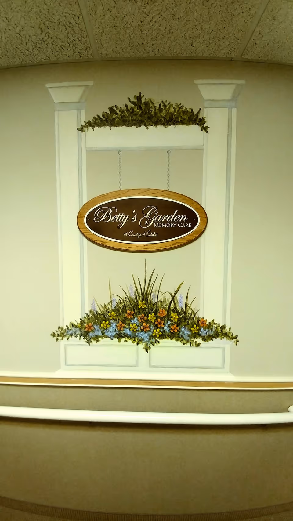 A hallway wall mural showing a window box of flowers and a hanging oval sign that reads "Betty's Garden Memory Care".