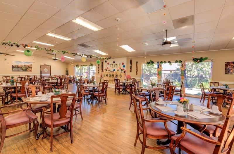 A spacious senior living dining room with multiple round wooden tables and chairs arranged neatly. Each table is set with white cups, plates, and napkins, and decorated with small flower arrangements. The room has large windows letting in natural light, a wooden floor, and ceiling lights. Colorful paper decorations hang from the ceiling and walls, creating a cheerful atmosphere.