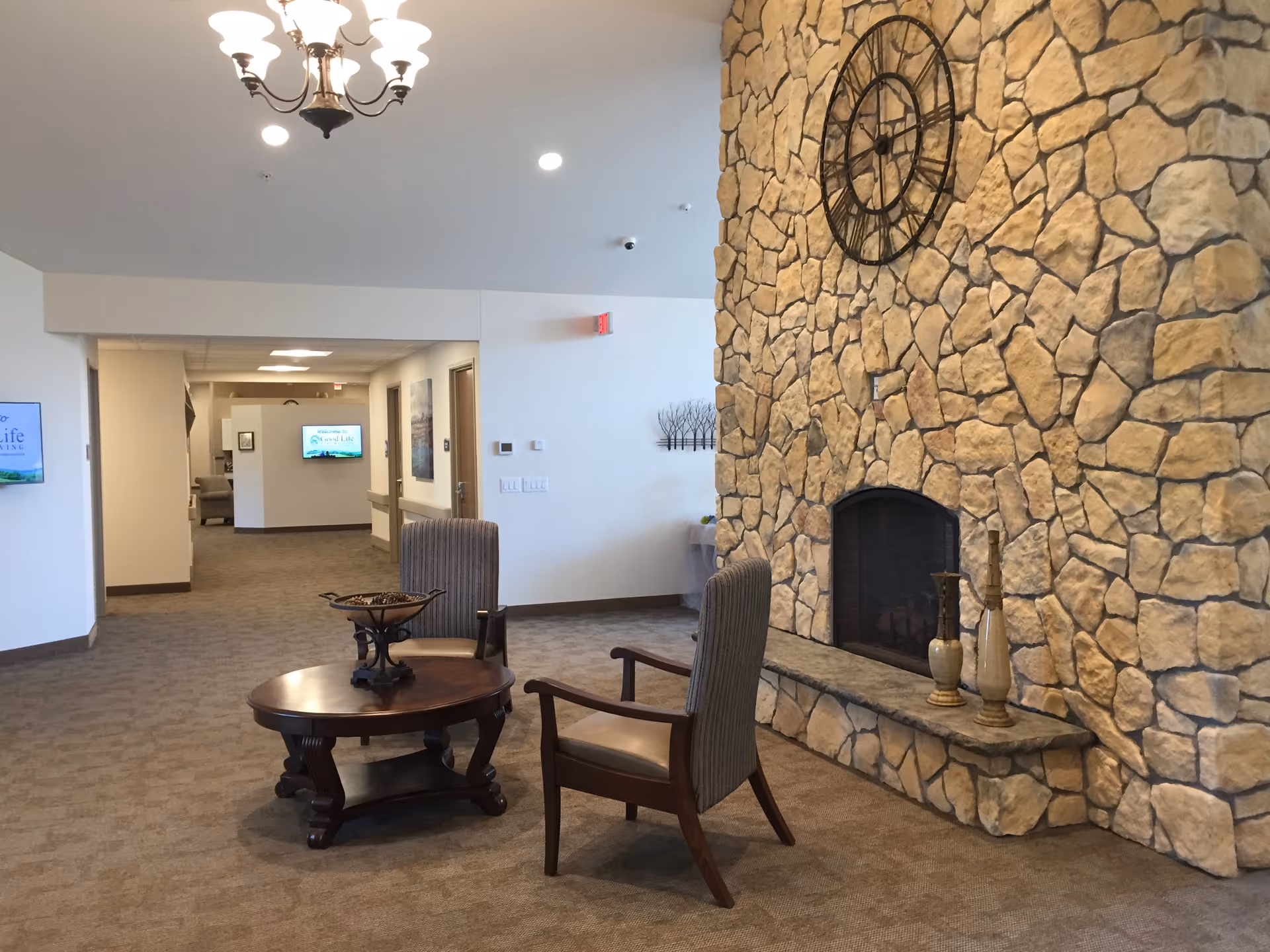 Lobby-style seating area with a stone fireplace, chairs, a coffee table, and a large wall clock in a senior living facility.