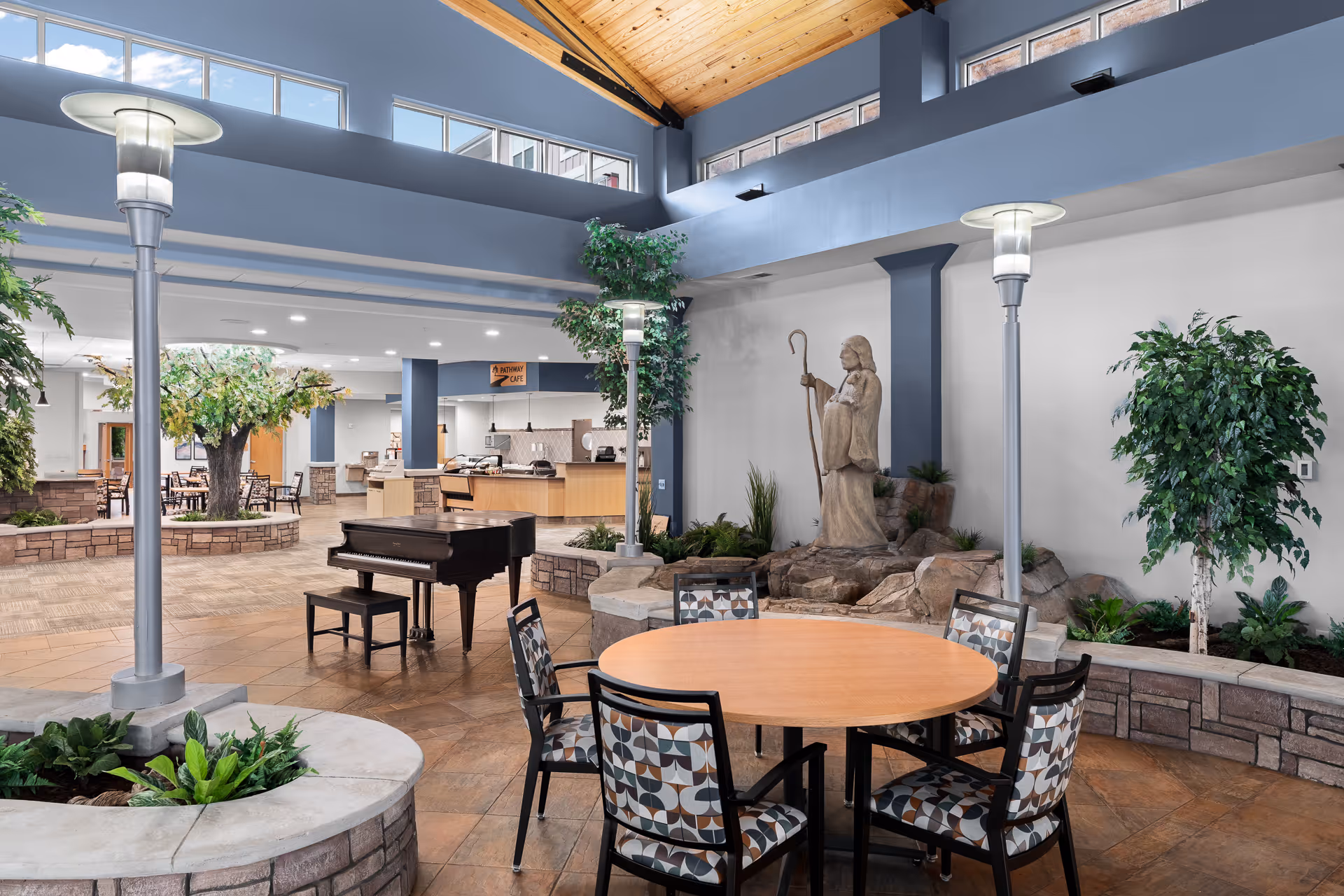 Atrium-style common area with a round table and chairs, a grand piano, plants, and a statue by a rock water feature under skylights.