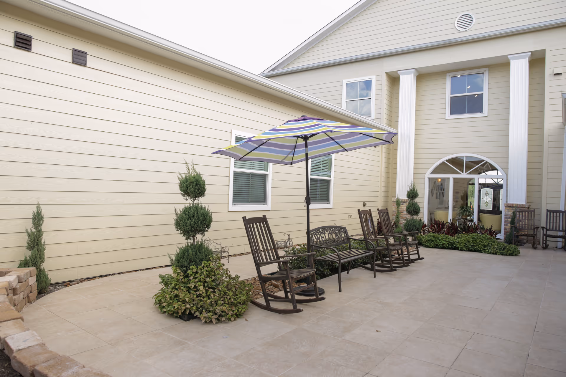 Courtyard patio with rocking chairs, a striped umbrella, potted plants and the entrance to a beige senior living building.