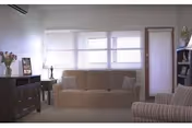 A bright living room with a beige sofa and a striped armchair. The room has large windows with white blinds, a dark wooden side table with a lamp, a cabinet with flowers and picture frames, and a bookshelf with various items.