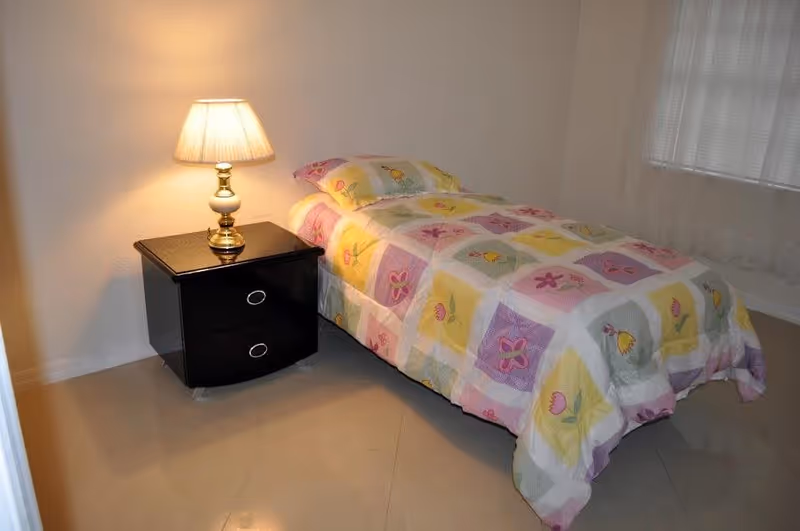 A small bedroom with a single bed covered in a colorful floral quilt. Next to the bed is a black nightstand with two drawers and a brass table lamp with a beige lampshade. A window with sheer white curtains is visible on the right side of the room.