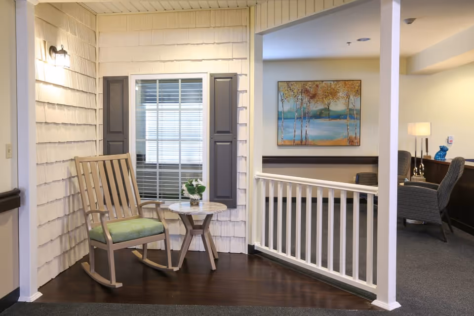 A cozy corner in a senior living facility featuring a wooden rocking chair with a green cushion next to a small round table with a potted plant. The wall behind has white shingle siding and a window with dark shutters. Adjacent to this corner is an open area with a white railing, a framed painting of trees and a lake on the wall, and a seating area with armchairs and a lamp.