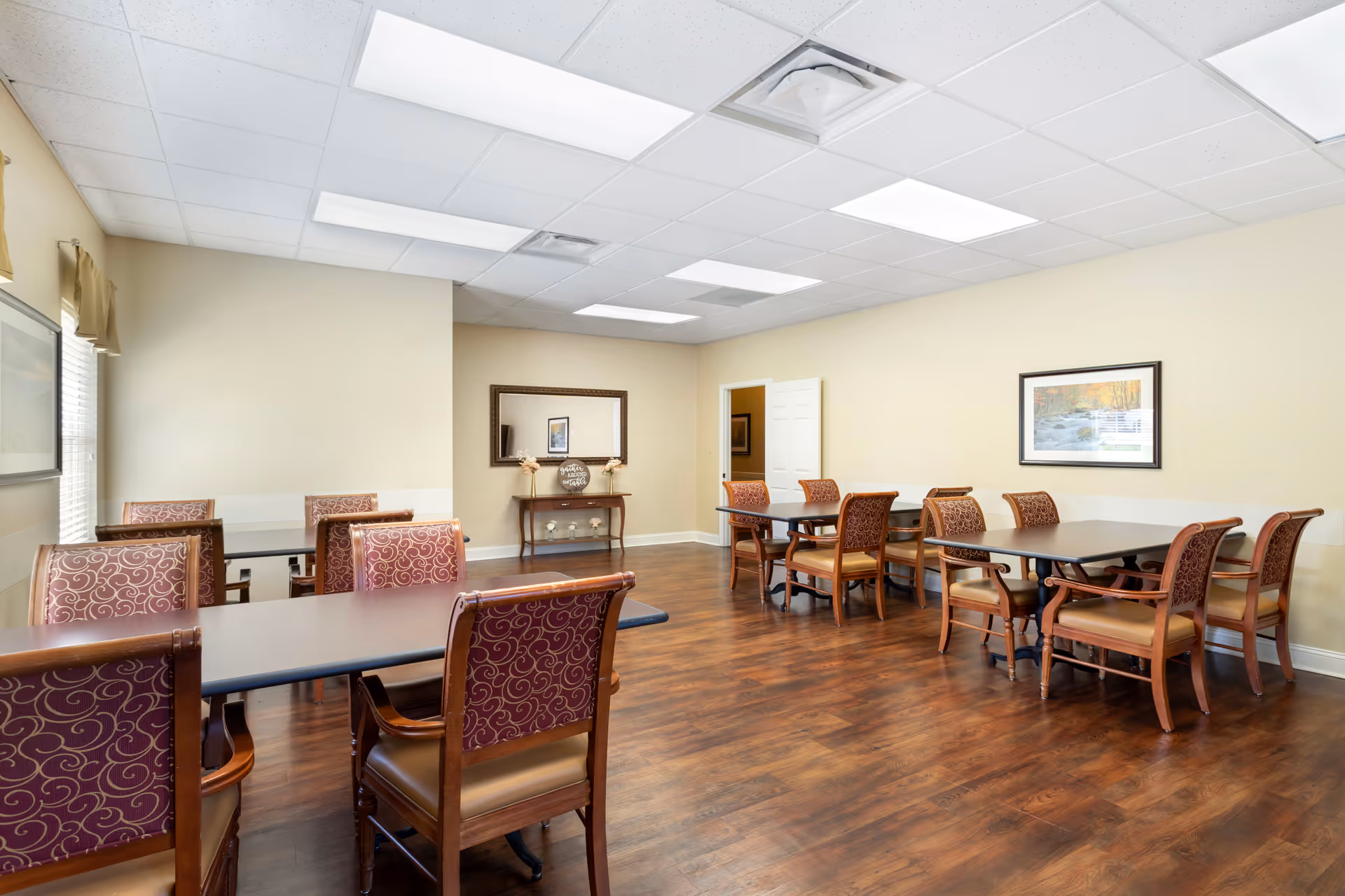 A well-lit dining room with multiple tables and chairs arranged neatly on a wooden floor. The walls are painted light beige and decorated with framed artwork and a large mirror above a small console table. The ceiling has recessed lighting panels.