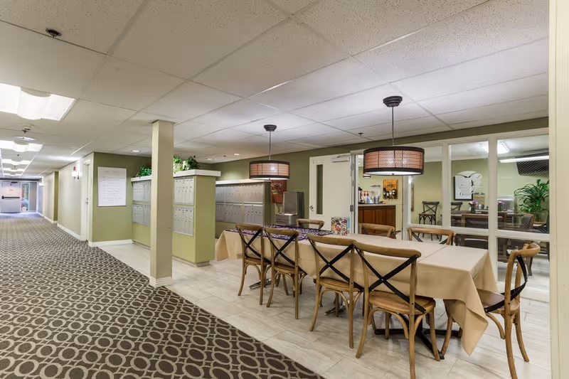 Interior view of a senior living facility hallway with a long table covered with a beige tablecloth and surrounded by wooden chairs. Two pendant lights hang above the table. On the left side, there are multiple mailboxes mounted on the wall. The hallway has patterned carpet and light green walls. A glass-walled room with additional seating and plants is visible in the background.