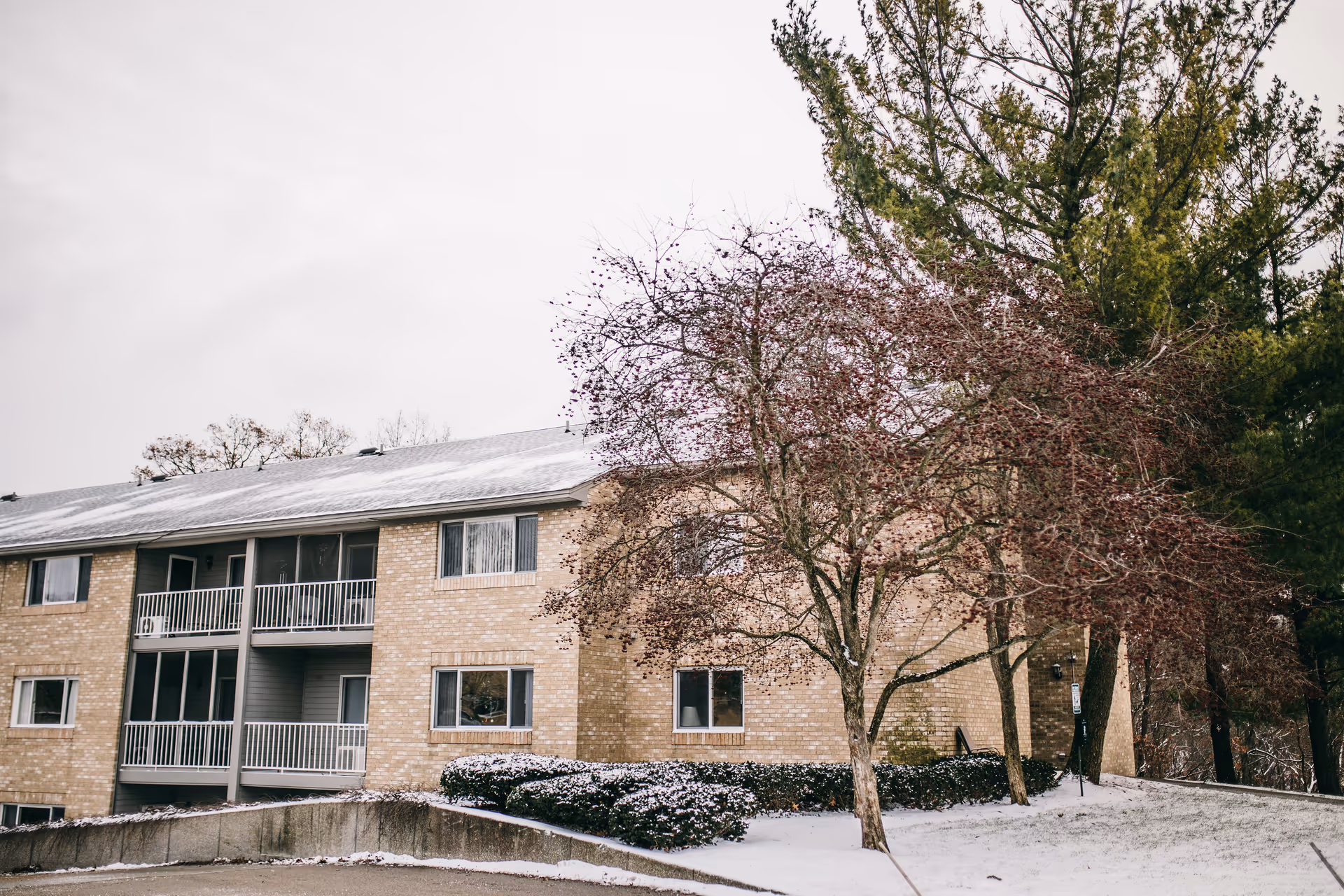 Front of a brick multi-unit retirement building with balconies, bare trees and a light layer of snow.