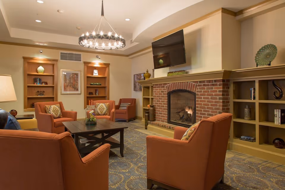 A cozy living room area with four orange armchairs arranged around a dark wooden coffee table. The room features a brick fireplace with a fire burning, a mounted flat-screen TV above it, built-in shelves with decorative items and books, and a modern chandelier hanging from the ceiling. The carpet has a patterned design, and the walls are painted in warm neutral tones.