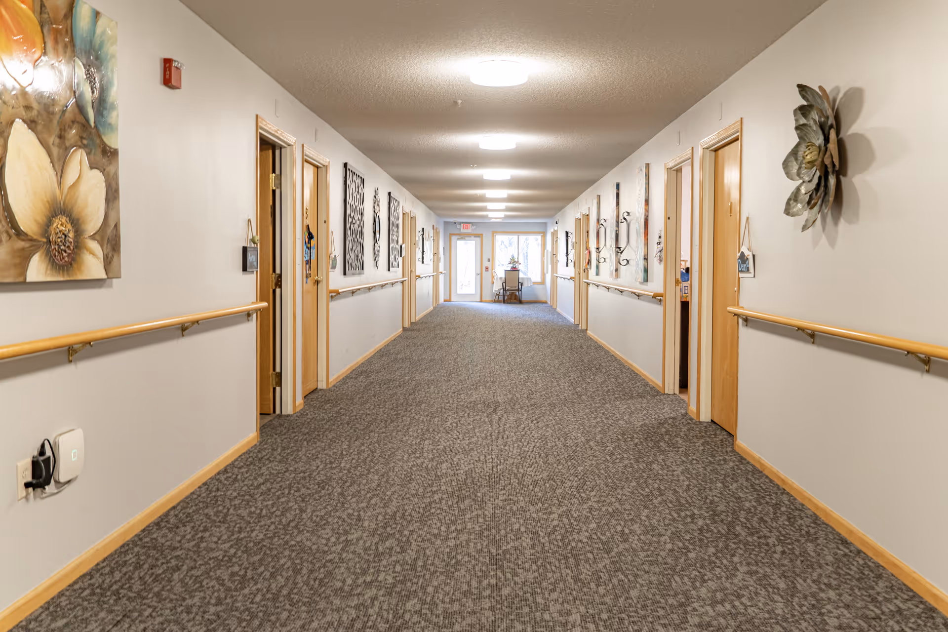 A long, carpeted hallway in a senior living facility with handrails on both sides and several wooden doors leading to rooms. The walls are decorated with various floral and abstract wall art. At the end of the hallway, there is a table with chairs near a window and a door with an exit sign above it.