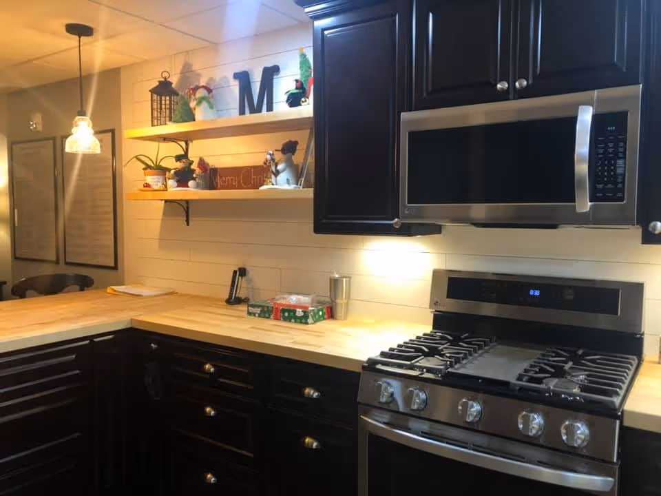 Modern kitchen with black cabinets, a stainless steel microwave and gas stove, wooden countertops, two wooden floating shelves with Christmas decorations, and a hanging pendant light over a corner counter.