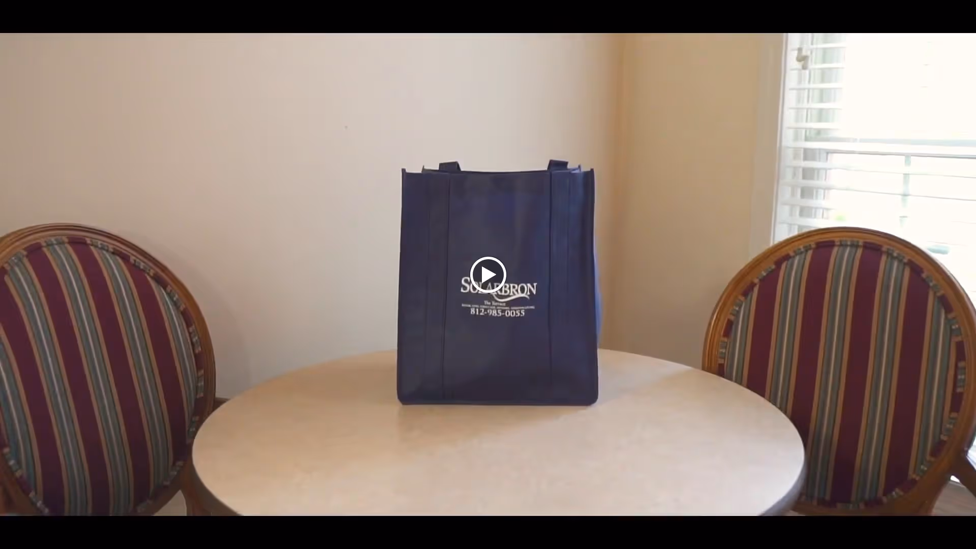 A round table with a beige surface is positioned between two wooden chairs with striped upholstery in shades of red, green, and beige. On the table, there is a dark blue tote bag with the text 'SOLARBRON Family-first Senior Living 812-985-0055' printed on it. A window with white blinds is visible in the background on the right side.