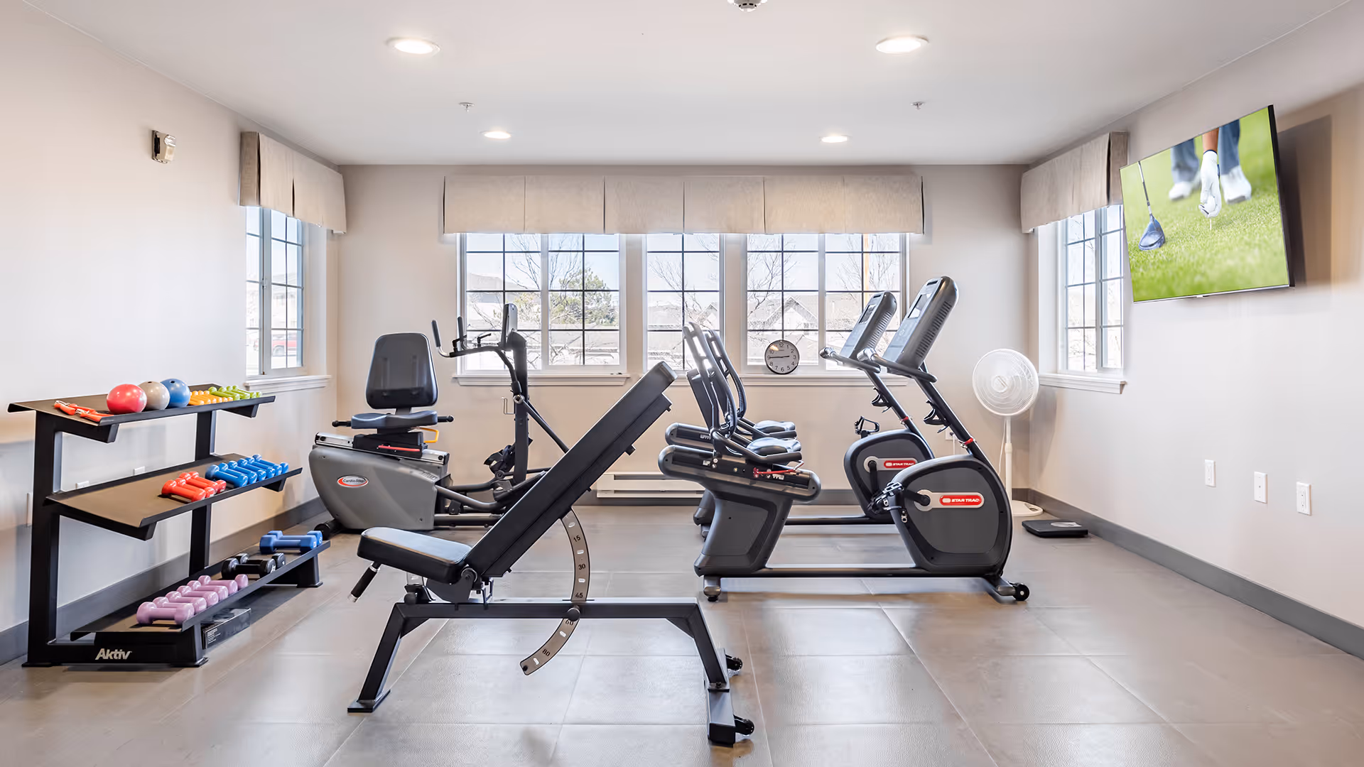 A bright fitness room with a weight bench and dumbbell rack in front of recumbent exercise bikes and a wall-mounted TV.