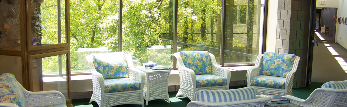 A bright indoor seating area with white wicker chairs and sofas featuring blue floral and striped cushions. The space has large floor-to-ceiling windows showing green trees outside, and a green carpet covers the floor. There is a small white wicker side table with books and tissues on it. A hallway with framed pictures and an exit sign is visible in the background.