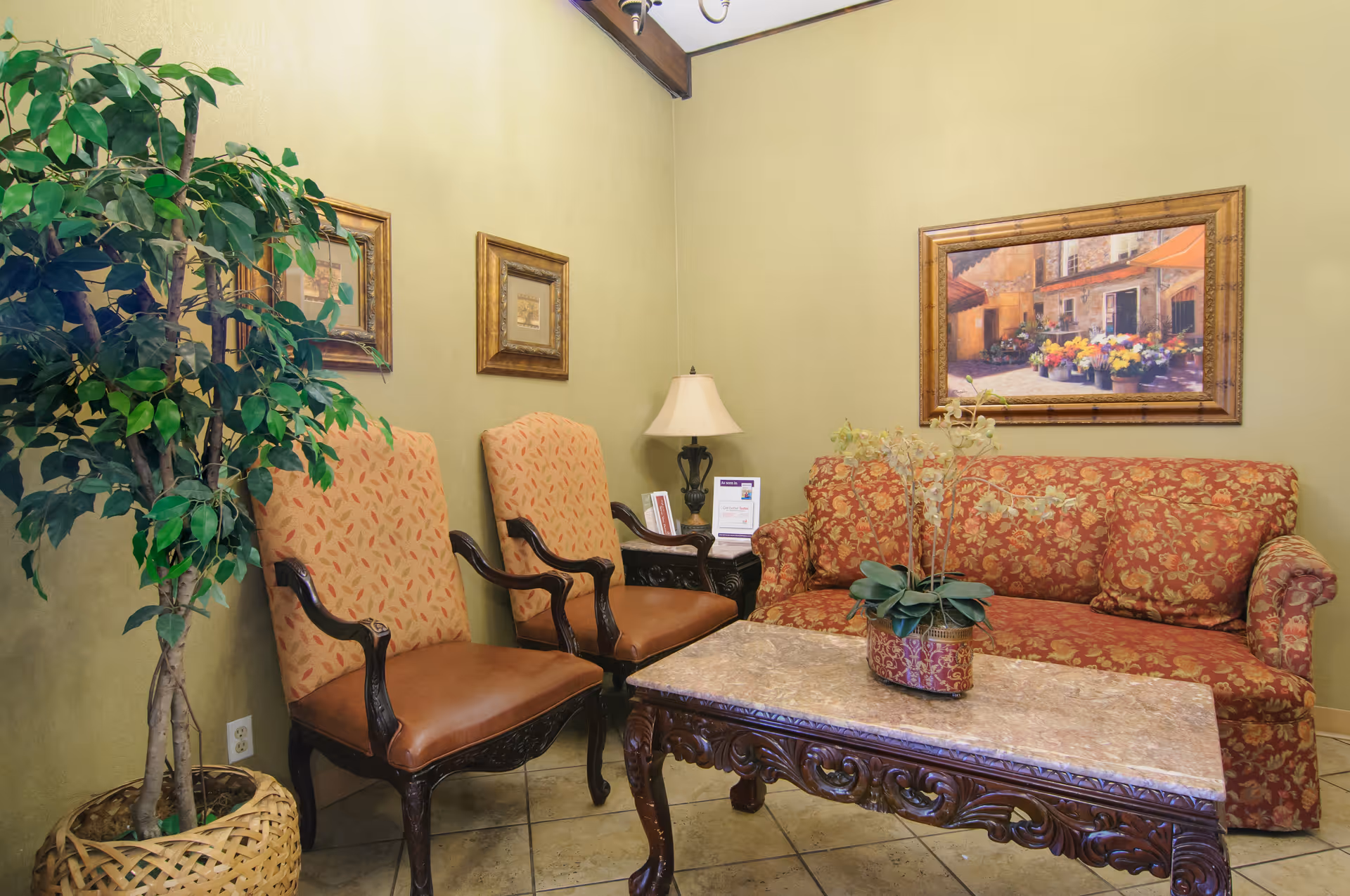 A cozy sitting area with two upholstered armchairs and a patterned sofa around a carved wooden coffee table with a plant on it. The walls are painted light green and decorated with framed artwork. A table lamp is placed on a small side table between the chairs and the sofa. There is a large potted plant in a woven basket on the left side.