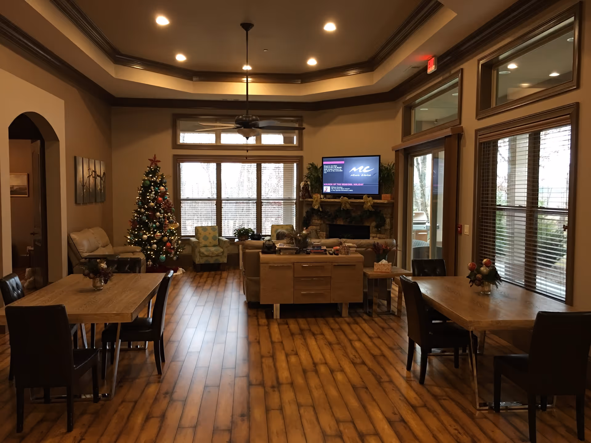 Interior view of a senior living community common area decorated for Christmas with a Christmas tree, wooden floors, two dining tables with chairs, a ceiling fan, large windows with blinds, a fireplace with a TV mounted above it, and comfortable seating including armchairs and a sofa.