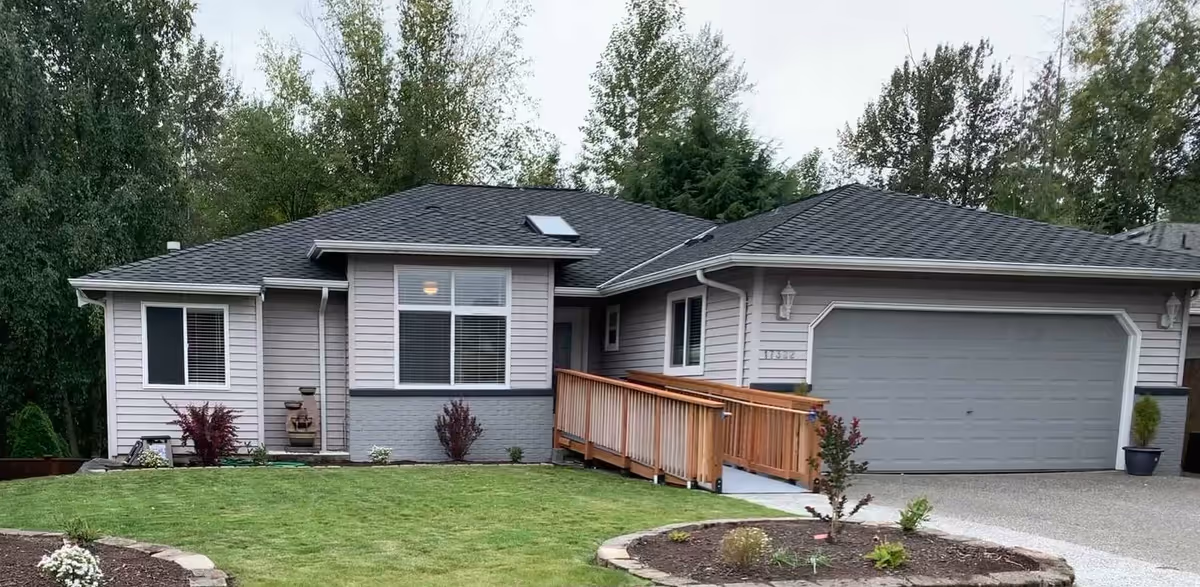 Single-story residential house with gray siding, a dark shingled roof, a wooden ramp leading to the front door, a two-car garage, and a well-maintained lawn with small shrubs and trees in the background.
