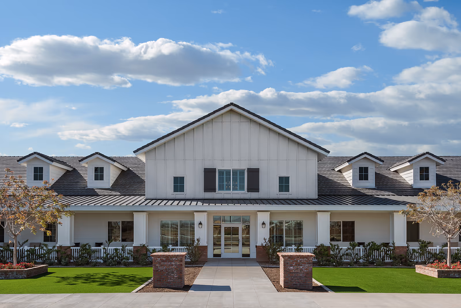 Front exterior of a white, ranch-style assisted living building with a covered entrance, porch and manicured lawn under a blue sky.