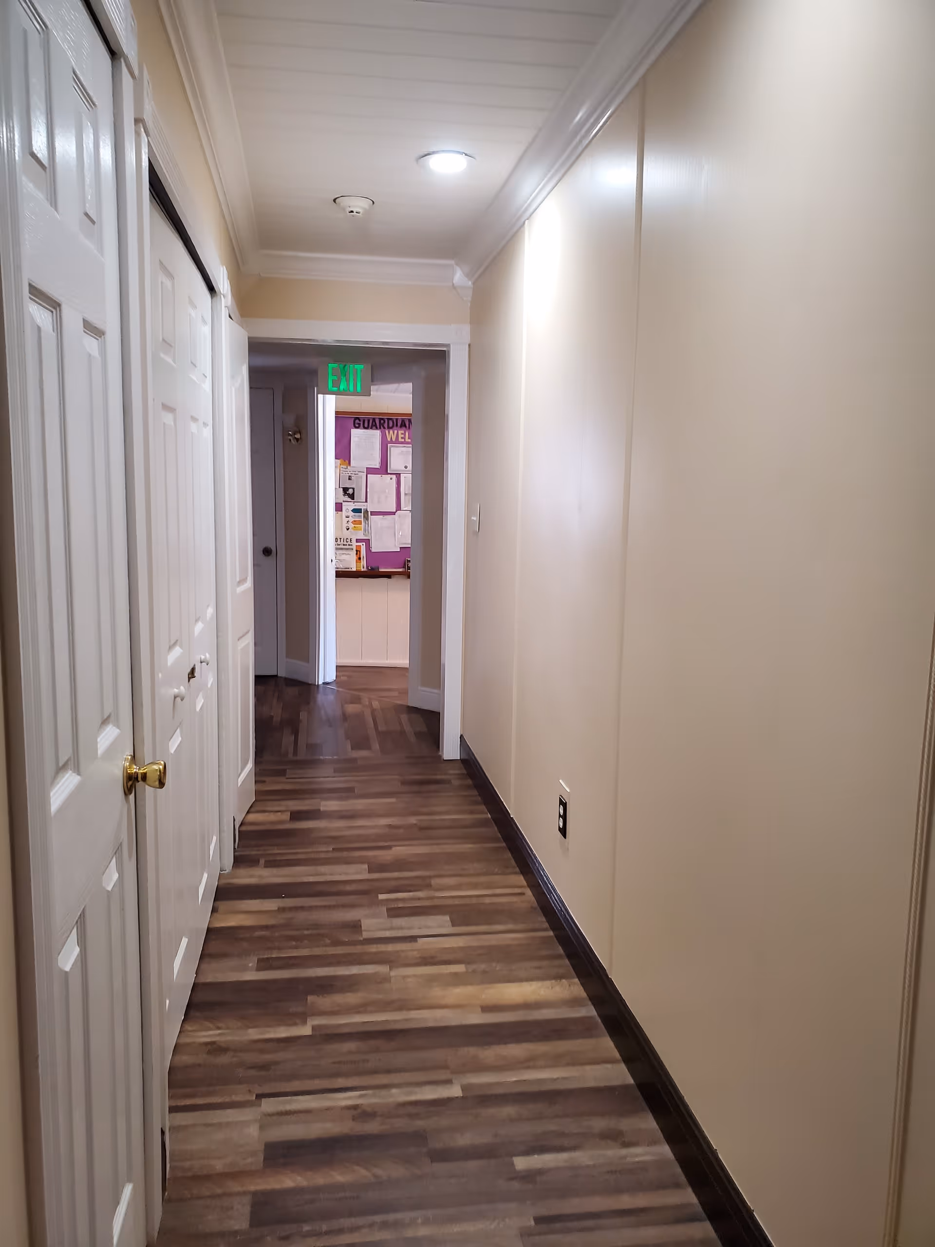 A narrow hallway with wood-patterned flooring and beige walls. On the left side, there are white doors with golden handles. The ceiling has recessed lighting and a smoke detector. At the end of the hallway, there is an open doorway leading to a room with a bulletin board and an illuminated green exit sign above the door.