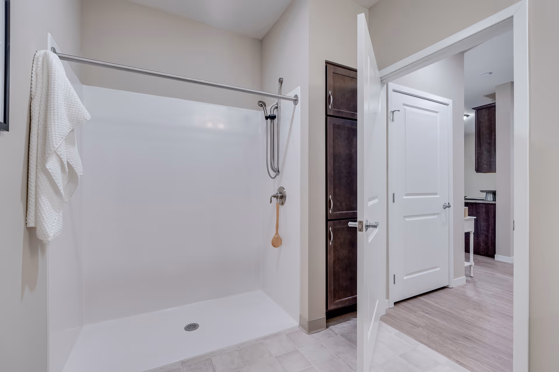A clean, modern bathroom with a white walk-in shower featuring a handheld showerhead and a towel hanging on a hook. The bathroom has light-colored walls and tile flooring. An open door leads to a hallway with wood flooring and dark wood cabinets.