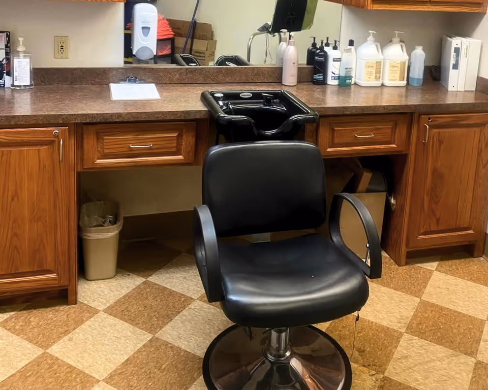 A salon station with a black salon chair in front of a countertop with a built-in black hair washing sink. The countertop has various bottles of hair care products and a large mirror behind it. The floor has a checkered pattern with brown and beige tiles, and there are wooden cabinets below the countertop.