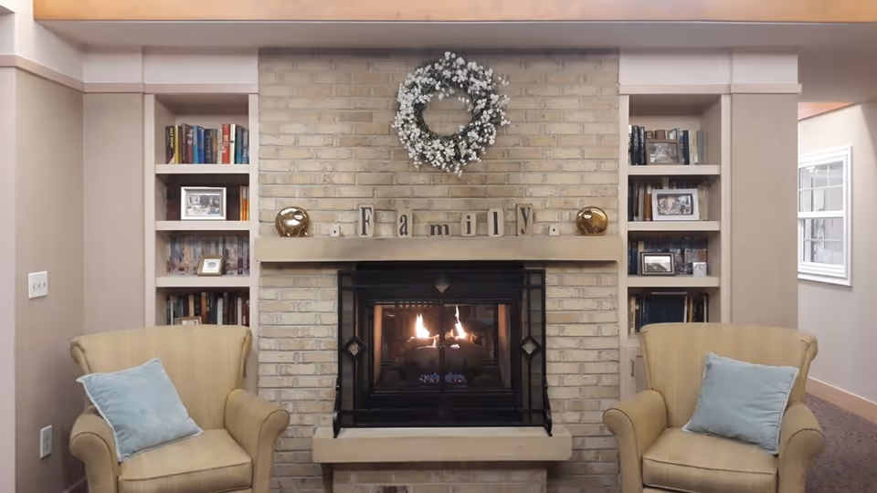 A cozy living room area with a lit fireplace set in a light brick wall. Above the fireplace is a decorative wreath and wooden blocks spelling out 'Family'. On either side of the fireplace are built-in bookshelves filled with books and framed photos. Two beige armchairs with light blue cushions are positioned facing the fireplace.