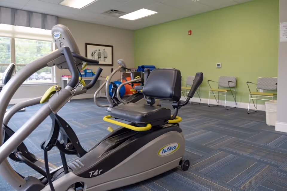 A fitness room with exercise equipment including a NuStep T4r recumbent cross trainer. The room has blue carpet tiles, light green walls, a large window letting in natural light, and several chairs lined up against the wall.