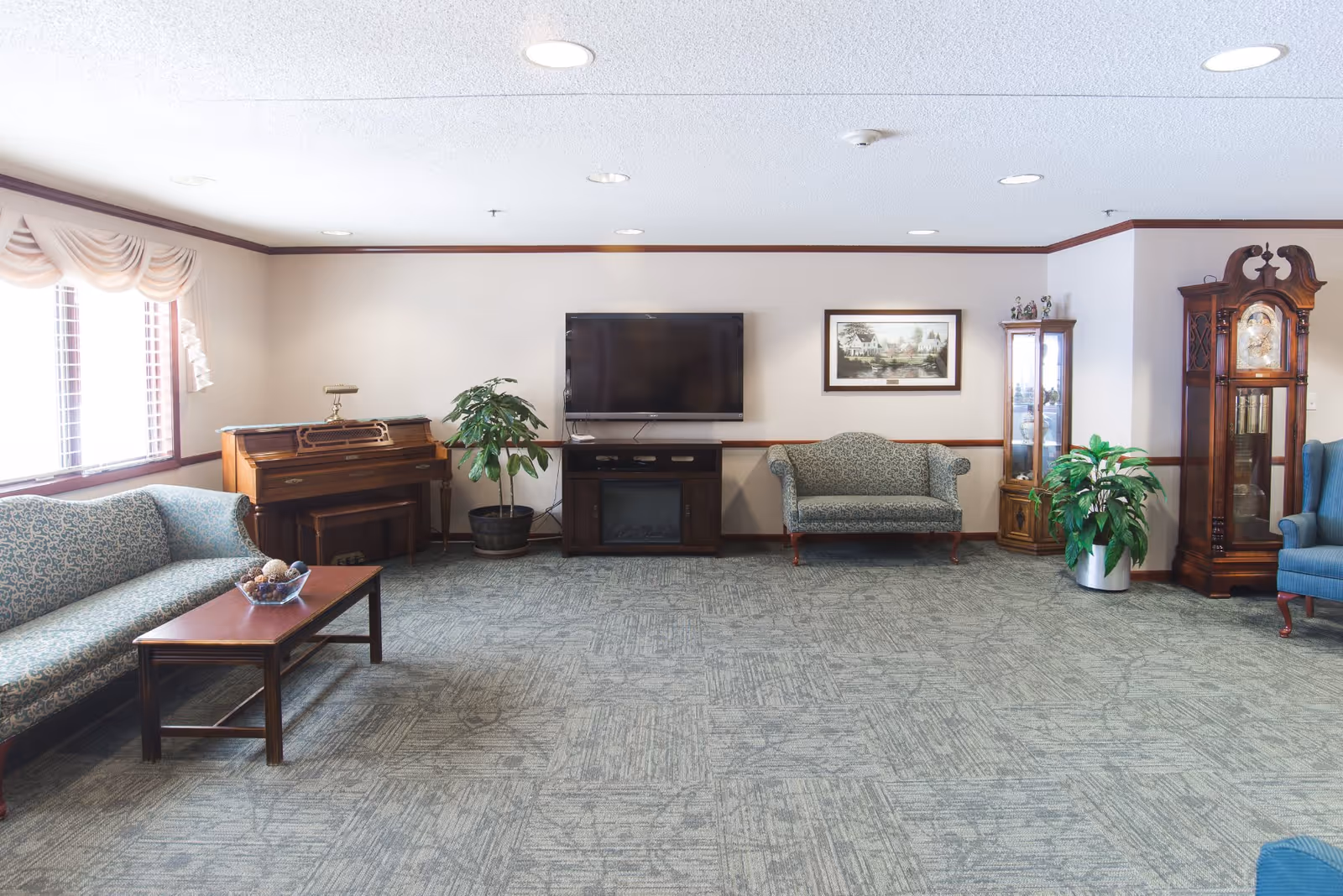 A spacious living room with patterned sofas, a wooden coffee table, a piano, a flat-screen TV mounted on the wall above a wooden cabinet, a framed picture, two potted plants, a glass display cabinet, and a tall grandfather clock. The room has large windows with draped curtains and carpeted flooring.