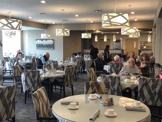 A dining room in a senior living facility with several round and rectangular tables covered with white tablecloths. The tables are set with cups, saucers, and napkins. Several elderly residents are seated at the tables, some eating and some conversing. The room has modern pendant lights hanging from the ceiling and large windows letting in natural light.