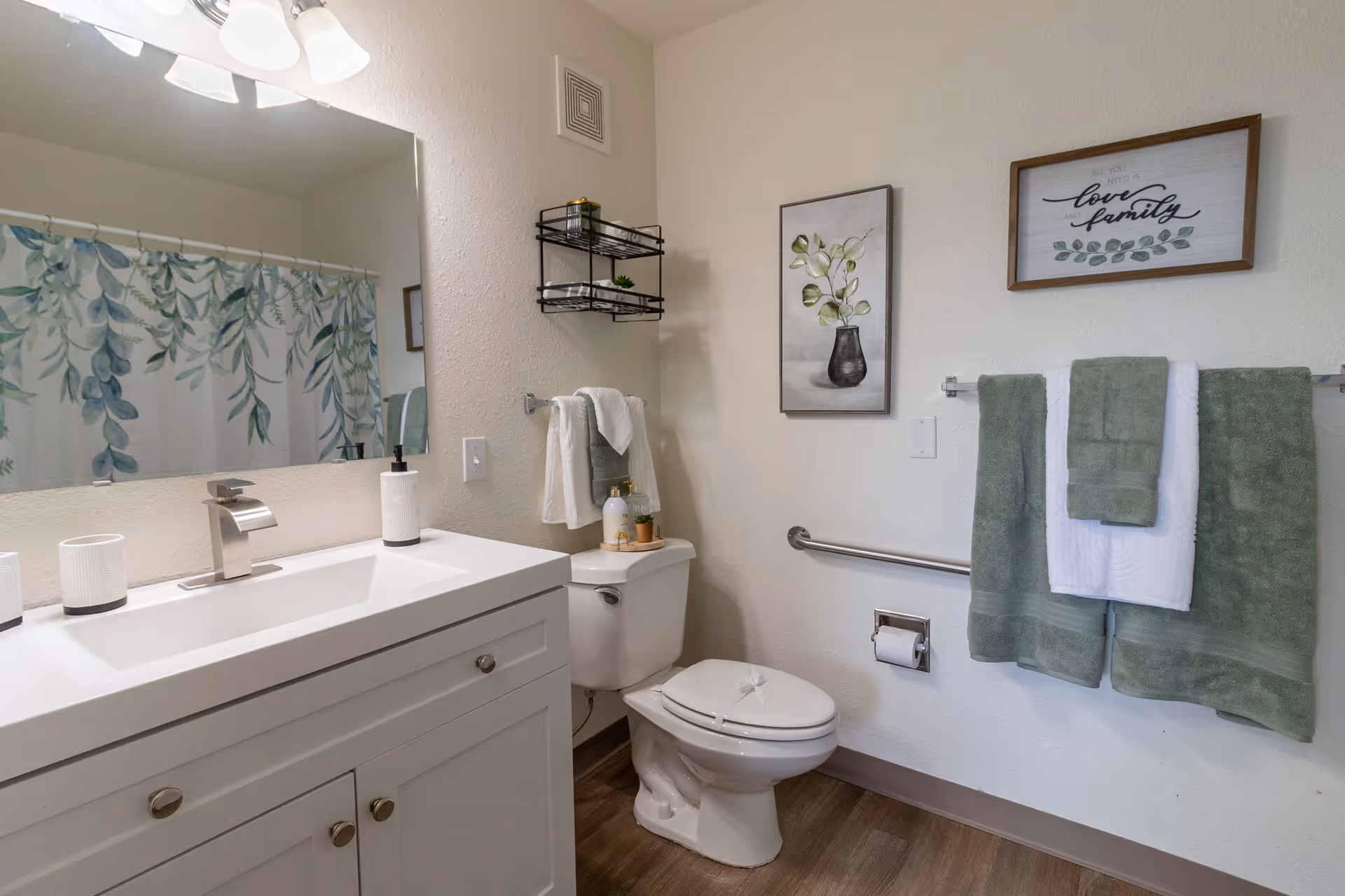 A clean and well-lit bathroom featuring a white vanity with a modern faucet, a large mirror, a toilet with a grab bar beside it, and a shower curtain with a green leafy pattern. The walls have two framed pictures, one with a plant and another with the words 'love family'. Green and white towels hang on a towel rack.