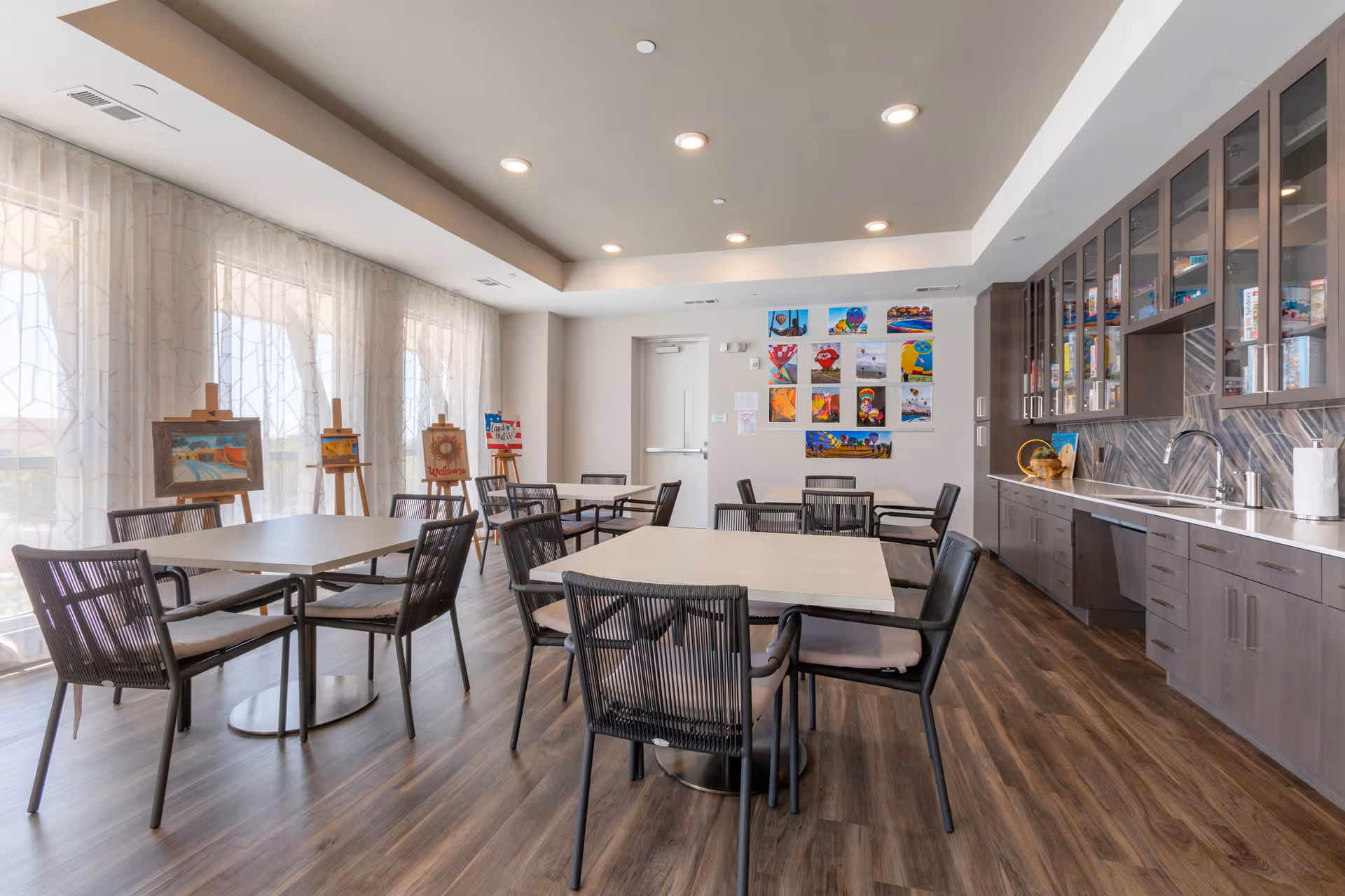 Bright communal dining and activity room with multiple tables and chairs, a kitchenette along the right wall, and artwork on easels and the back wall.
