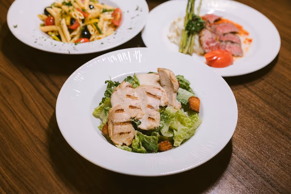 Three white plates of food on a wooden table. The closest plate contains a grilled chicken salad with lettuce and croutons. The plate in the background on the left has a pasta dish with cherry tomatoes and olives. The plate in the background on the right has sliced meat, mashed potatoes, asparagus, and a tomato garnish.