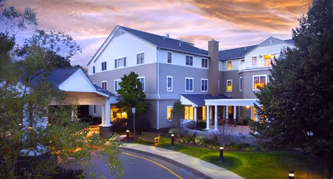 Exterior view of a multi-story senior living facility building at dusk with lights on inside. The building has a covered entrance with a driveway and landscaped greenery around it. The sky shows a colorful sunset with clouds.
