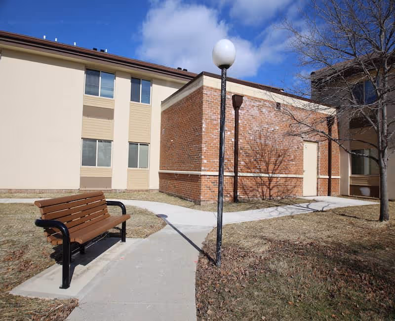 Outdoor courtyard with a bench and lamppost in front of a beige-and-brick multi-story building under a blue sky.