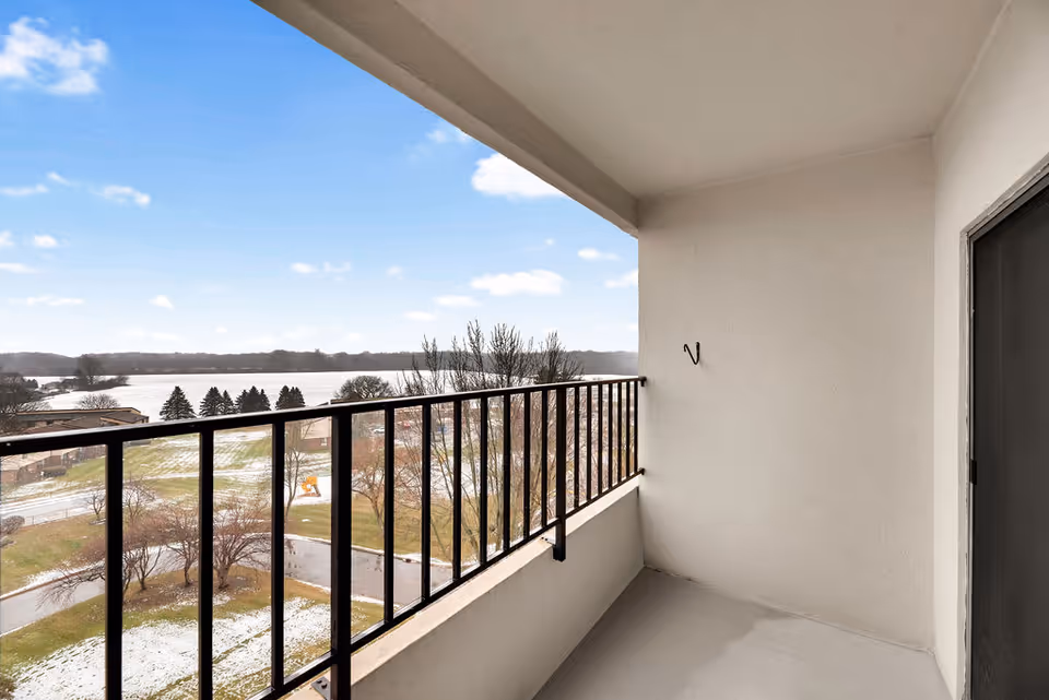 View from a covered balcony with black metal railing overlooking a landscape with trees, a road, and a body of water in the distance under a partly cloudy sky.