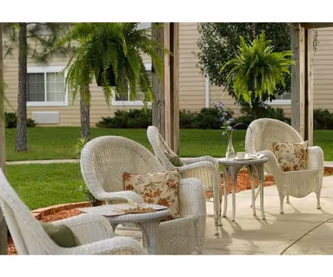 Outdoor seating area with white wicker chairs and small tables on a concrete patio, decorated with floral cushions and a vase with flowers, surrounded by green grass and hanging ferns under a wooden pergola.