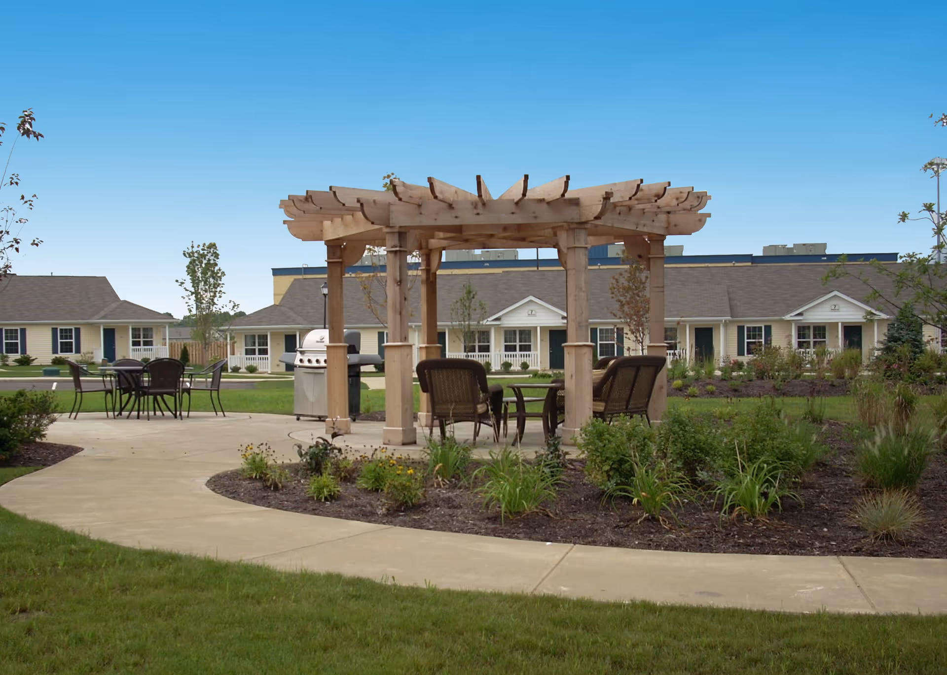 Outdoor seating area with a wooden pergola, chairs, and a table on a paved patio surrounded by landscaped garden beds and grass. In the background, there are single-story residential buildings under a clear blue sky.